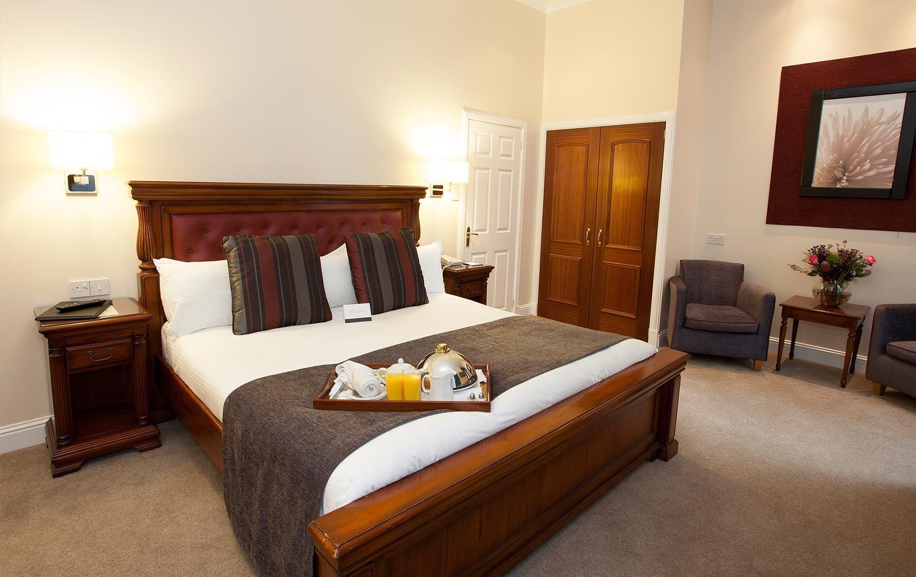 A hotel room with a large bed, a wooden wardrobe, a chair, a small table with a vase, and a tray with breakfast items.