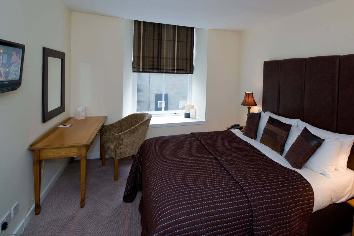 A hotel room with a bed, a desk, a chair, a window with a view, and a nightstand with a lamp.