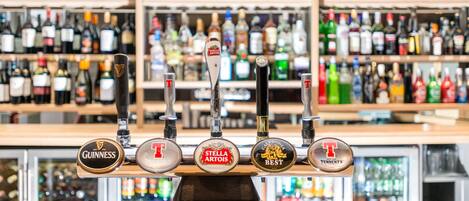 Um bar com várias torneiras, incluindo marcas como Guinness e Stella Artois, e prateleiras com diversas garrafas de bebidas ao fundo.