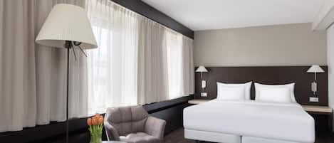 A modern hotel room with a large bed, a small round table with a vase of flowers, a chair, and a lamp.