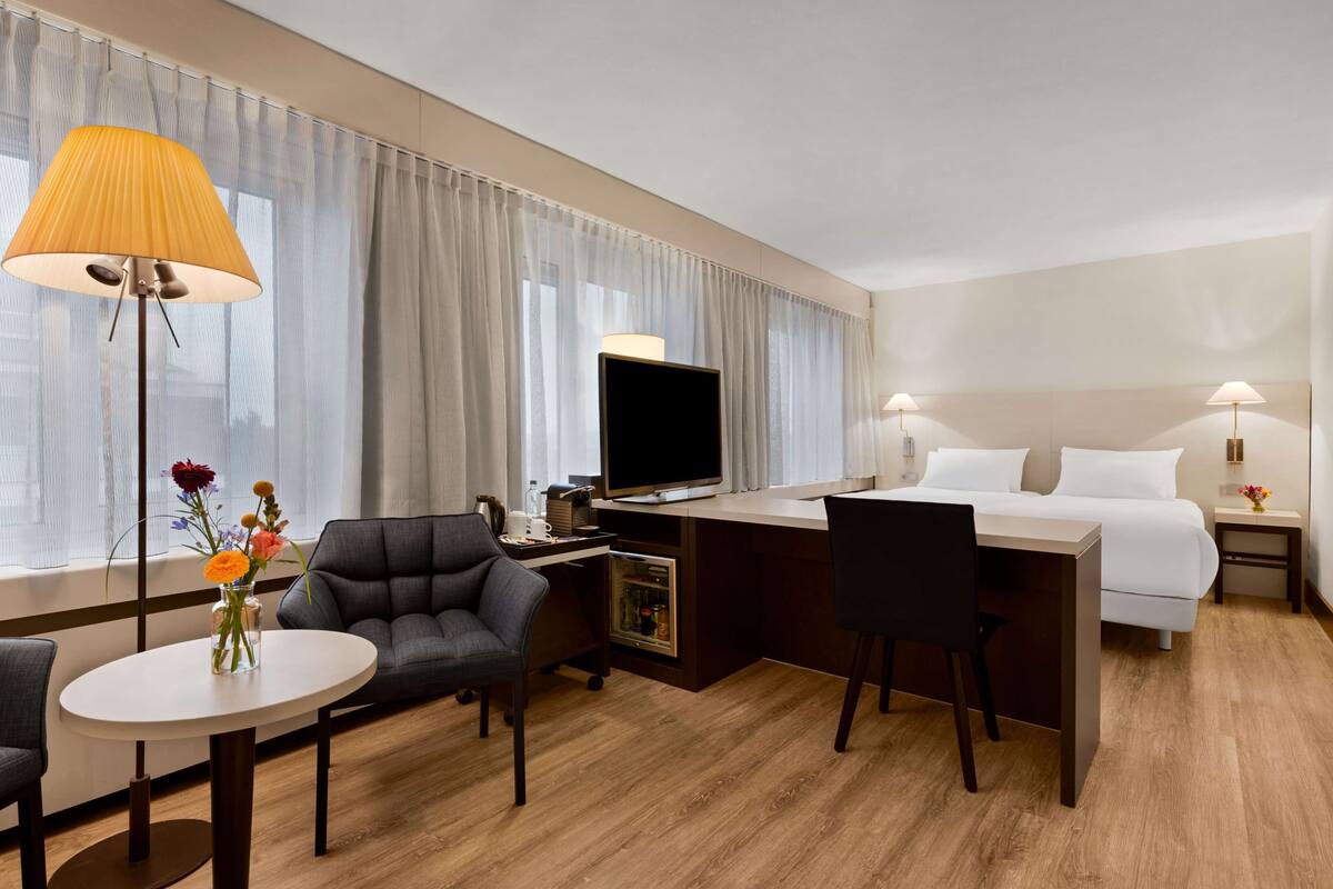 A hotel room with a bed, desk, chairs, a television, and a lamp.