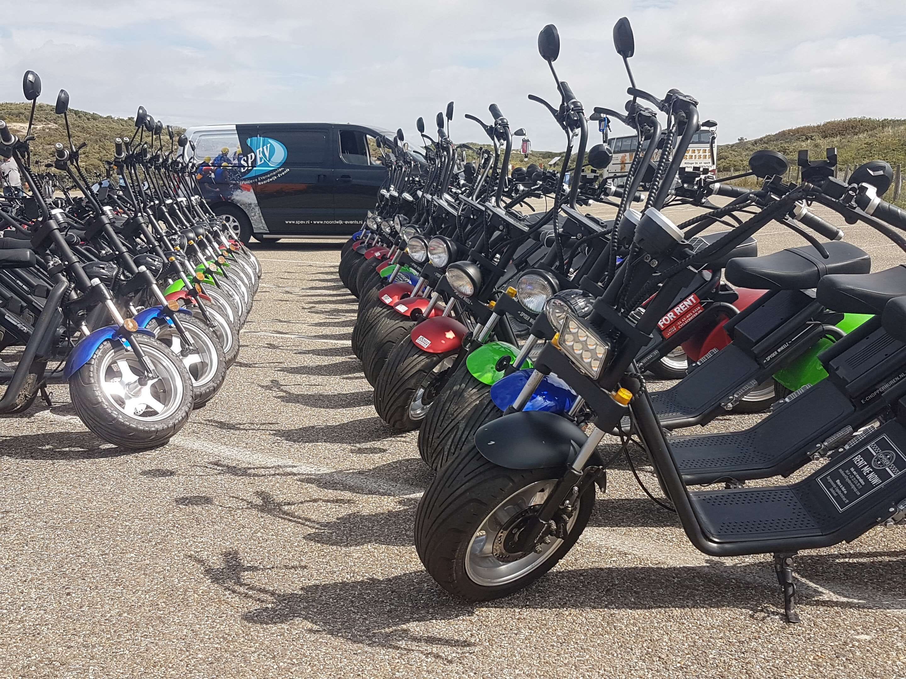 A row of electric scooters parked in a line, with a van in the background.