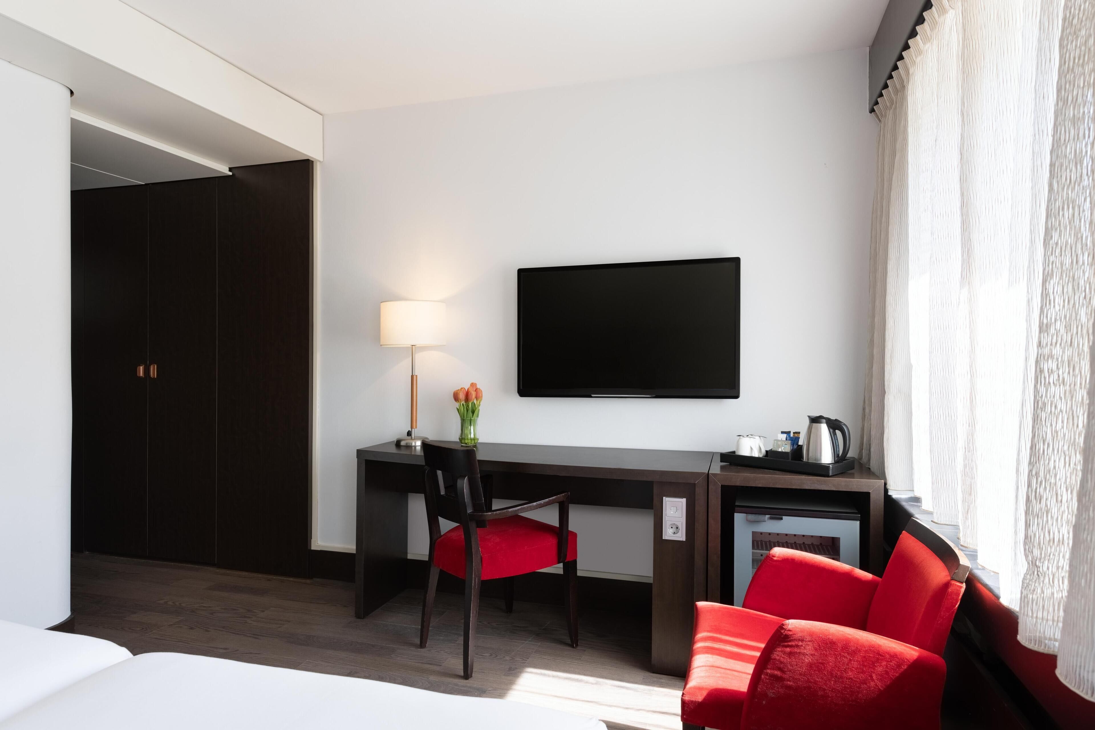 A hotel room with a bed, a desk with a chair, a TV, a small kitchenette, and a window with curtains.