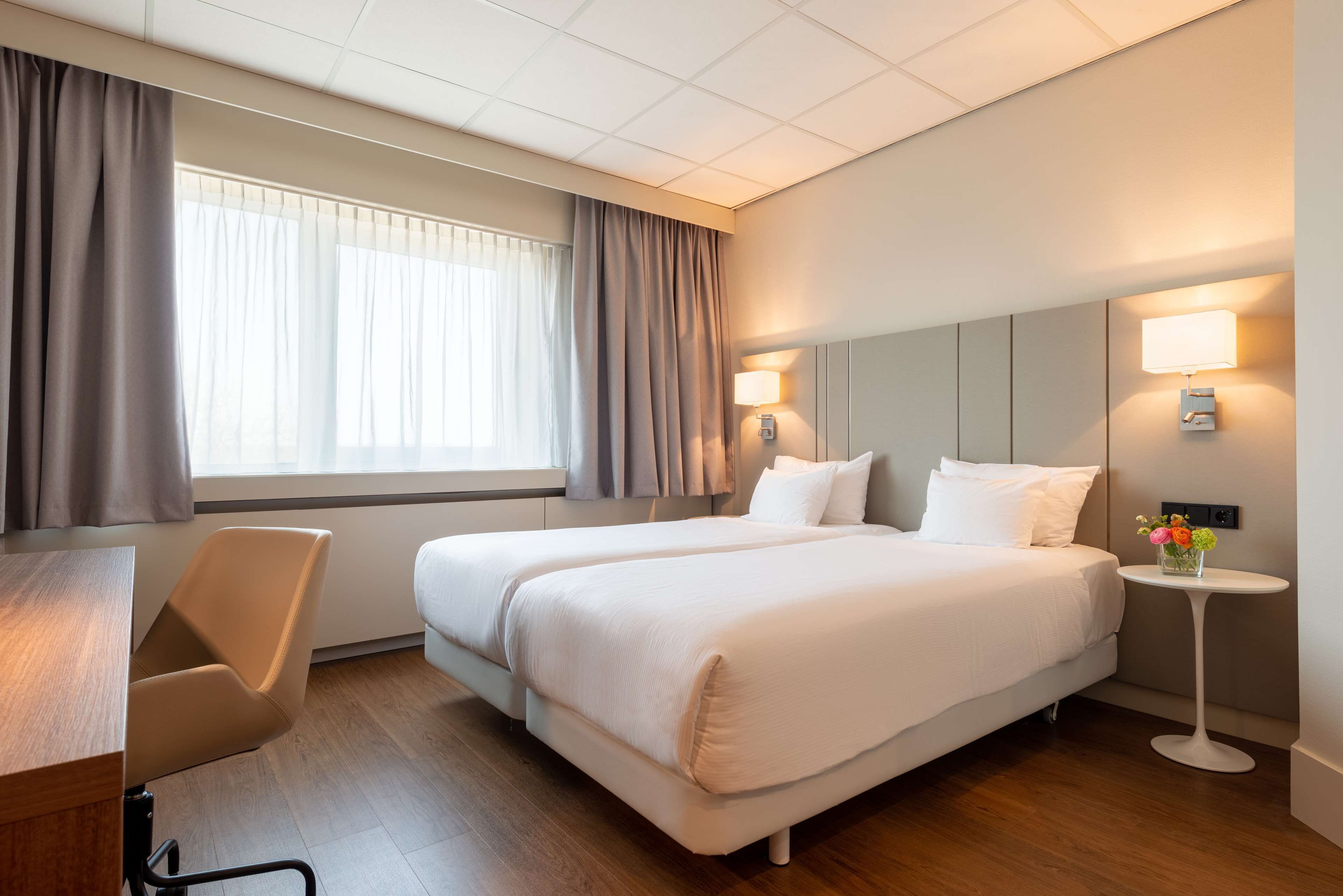 Habitación de hotel con dos camas, un escritorio y una pequeña mesa con lámpara y flores.