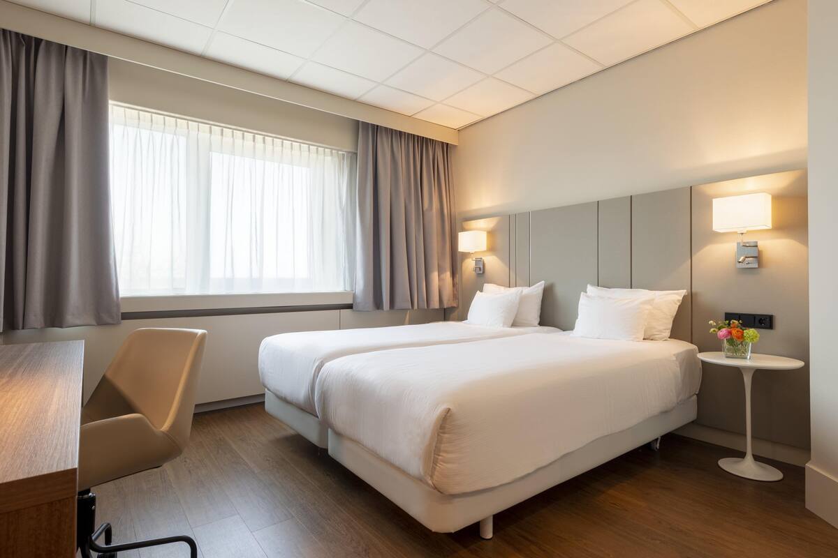 A hotel room with two beds, a desk, a chair, a small table with a lamp, and a window with curtains.