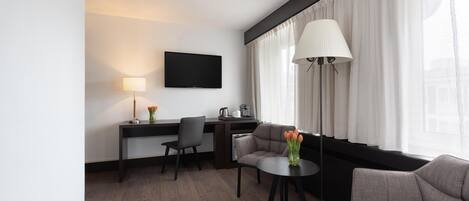 A modern hotel room with a flat-screen TV, a desk with a chair, a sofa, a small round table with a vase of flowers, and a large window with sheer curtains.