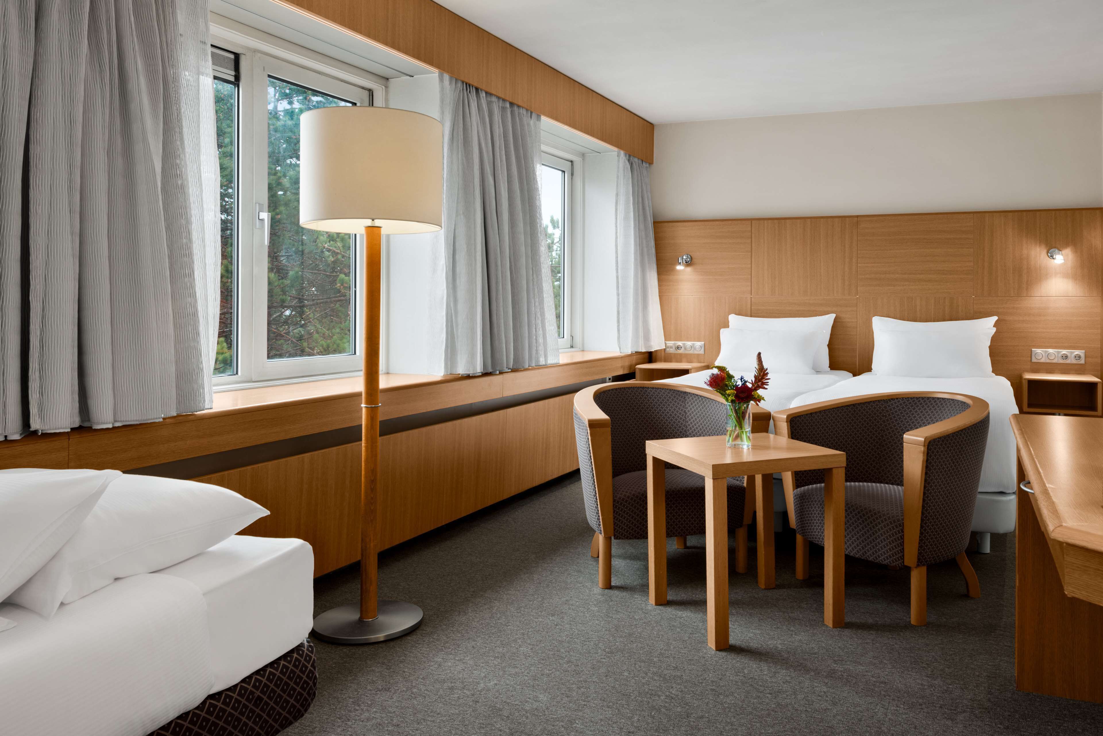 Une chambre d’hôtel avec un lit, deux chaises, une petite table, une lampe et une fenêtre avec des rideaux.