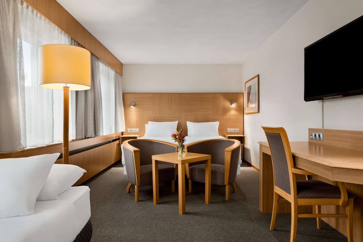 A hotel room with a bed, a desk, two chairs, a lamp, a TV, and a window with curtains.