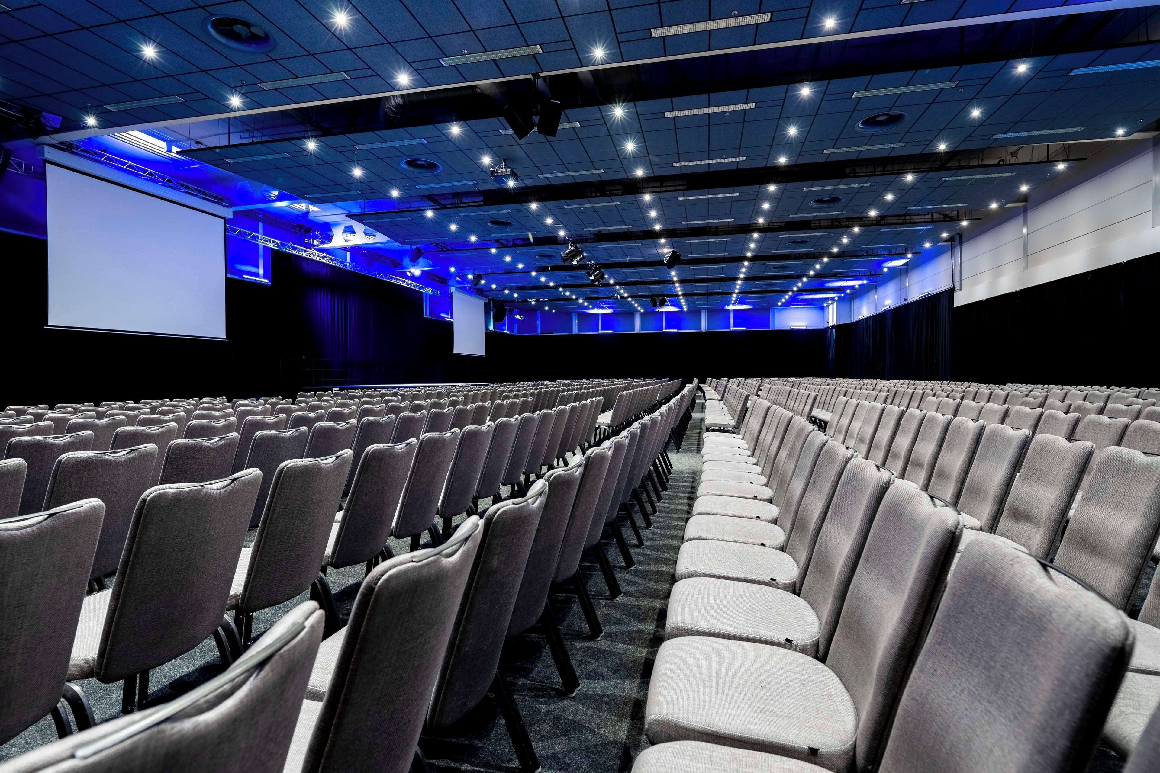 Un gran auditorio interior con filas de sillas grises y una pantalla grande al frente.