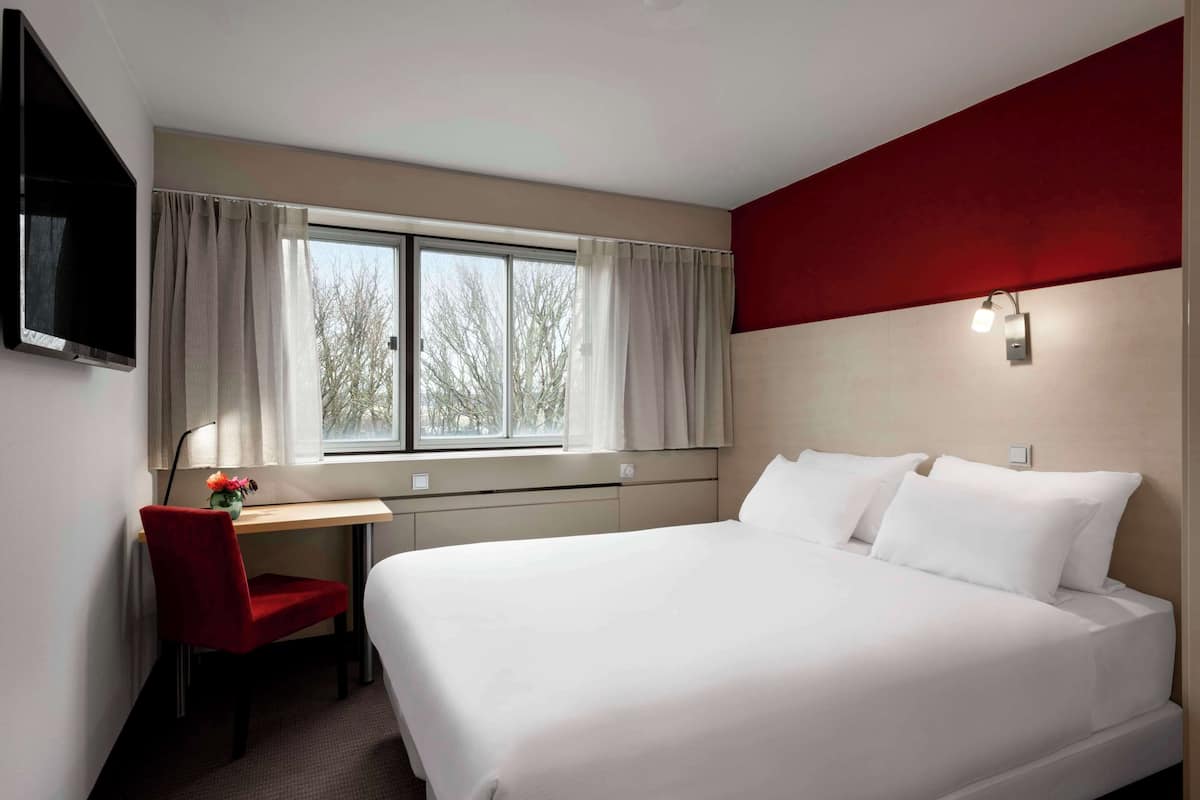 A hotel room with a bed, a desk, a chair, a TV, and a window with curtains.
