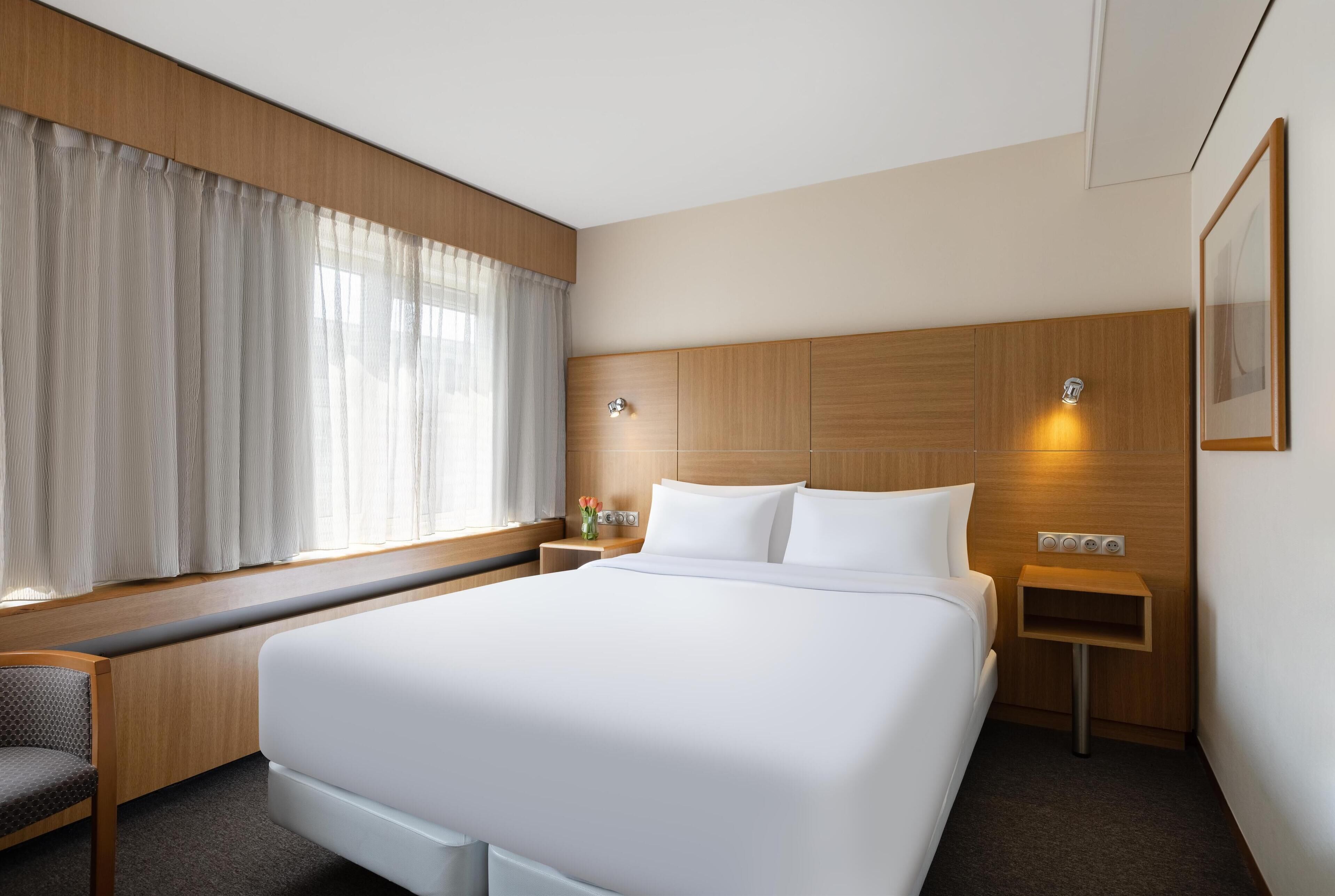 A hotel room with a large bed, a bedside table, a chair, a window with curtains, and a wall-mounted light.