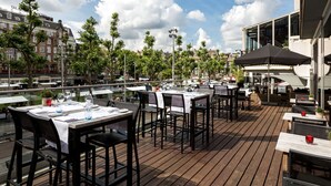 An outdoor dining area with tables and chairs, overlooking a canal with buildings in the background. - NH Amsterdam Caransa (Amsterdam)