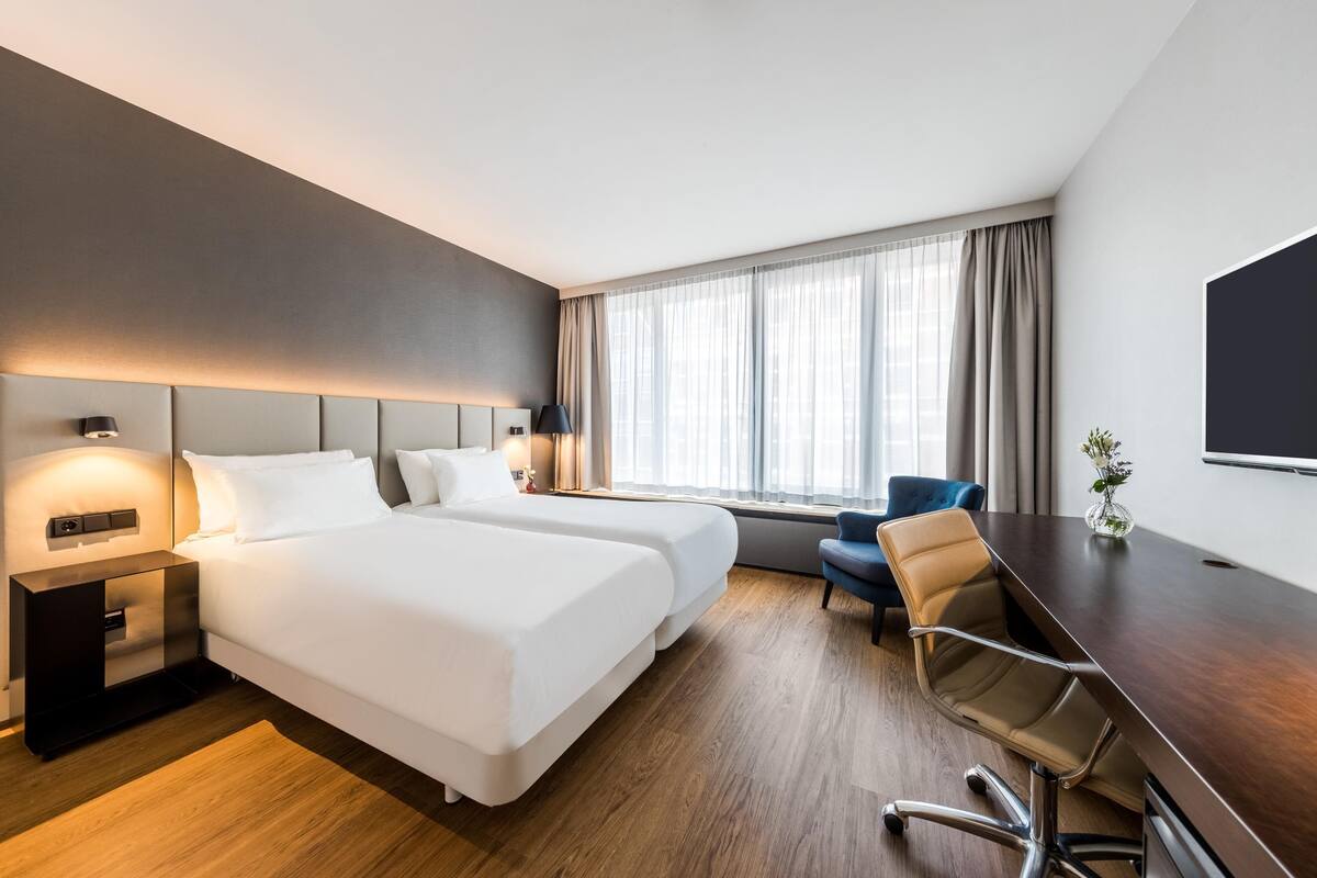 A hotel room with two beds, a desk, a chair, a TV, and a window with curtains.