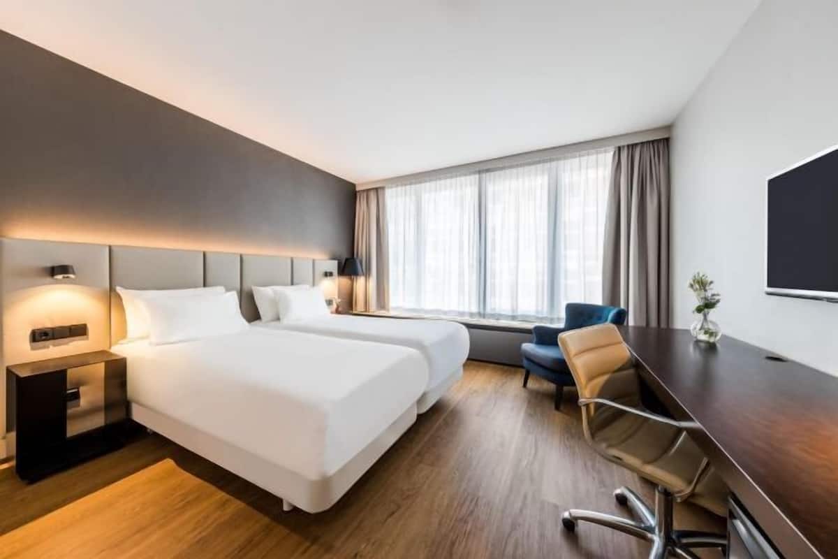 A hotel room with two beds, a desk, a chair, a TV, and a window with curtains.