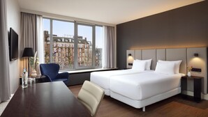 A hotel room with a large window, a bed, a desk, and a chair. - NH Amsterdam Caransa (Amsterdam)