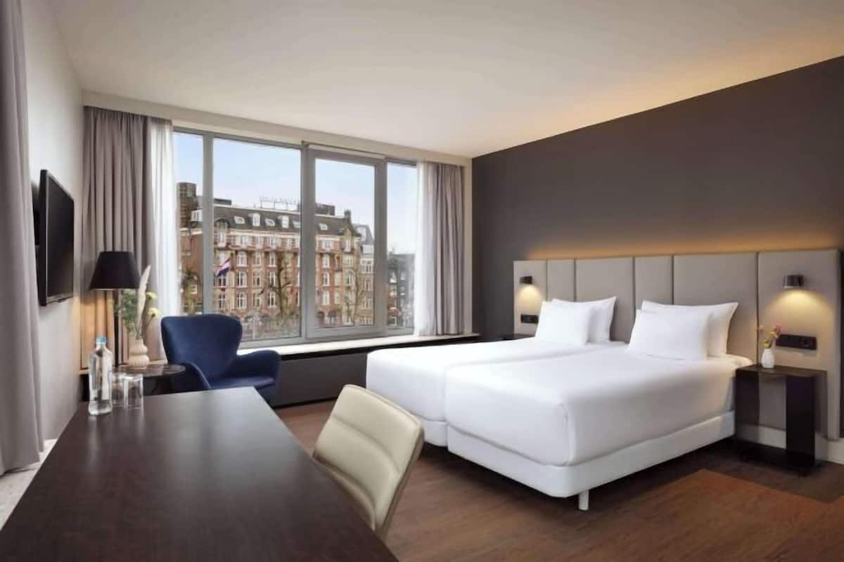 A modern hotel room with a large bed, a desk, and a chair. There is a window with a view of buildings outside.