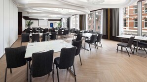 A spacious conference room with multiple tables and chairs, large windows offering a view of the city, and a modern chandelier. - NH Amsterdam Caransa (Amsterdam)
