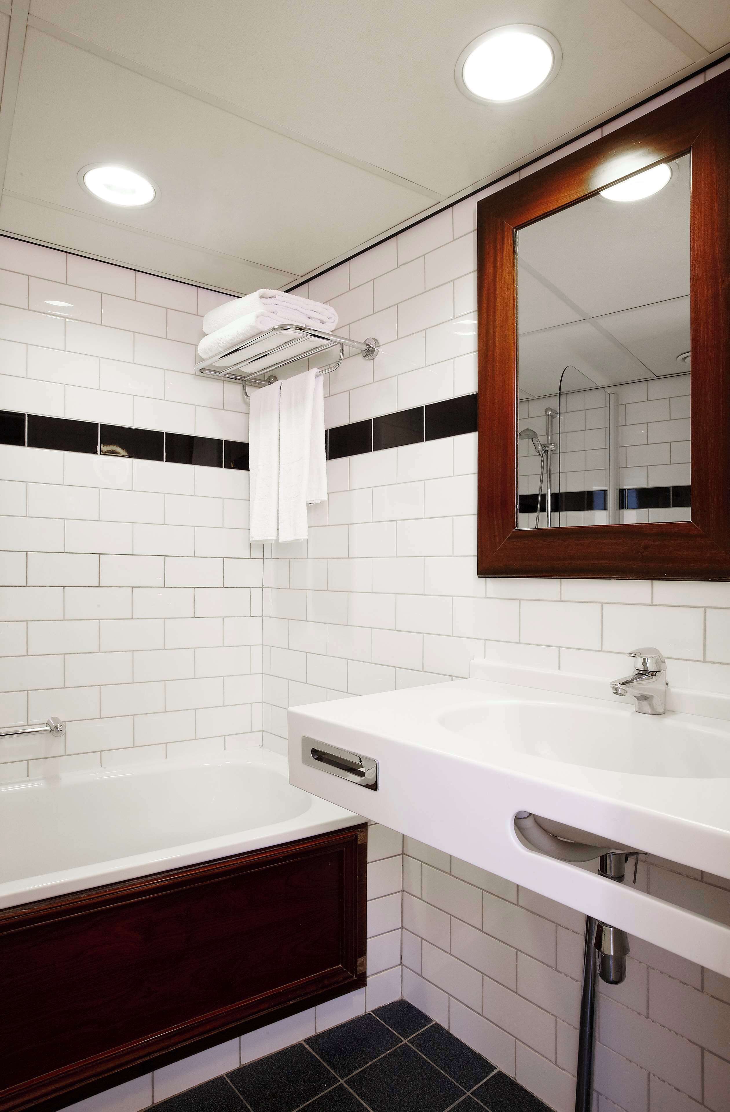 A modern bathroom with white tiles, a wooden vanity, a large mirror, and a bathtub.