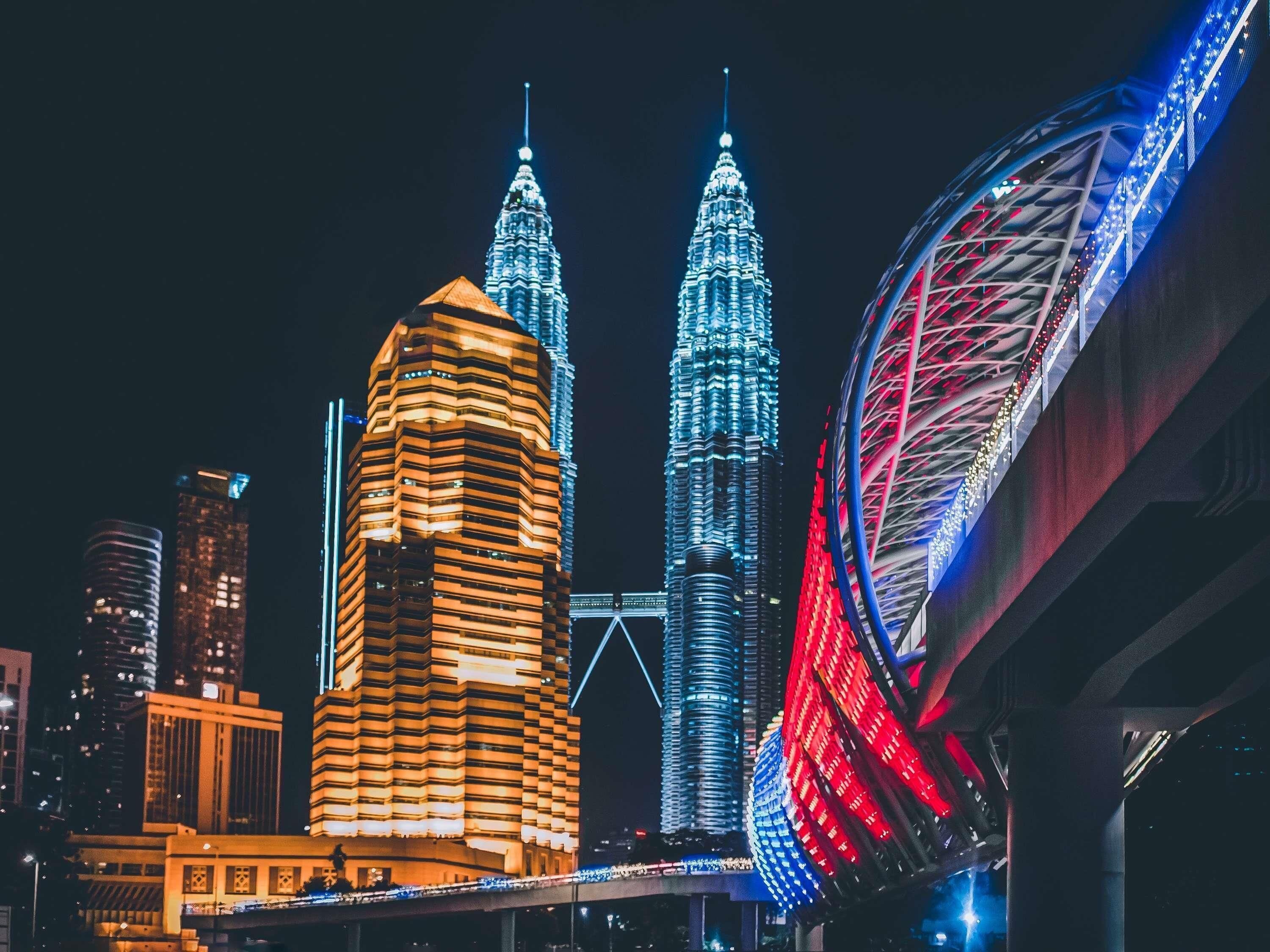 A cityscape at night with illuminated skyscrapers, including the Petronas Towers and a distinctive building with a unique facade.