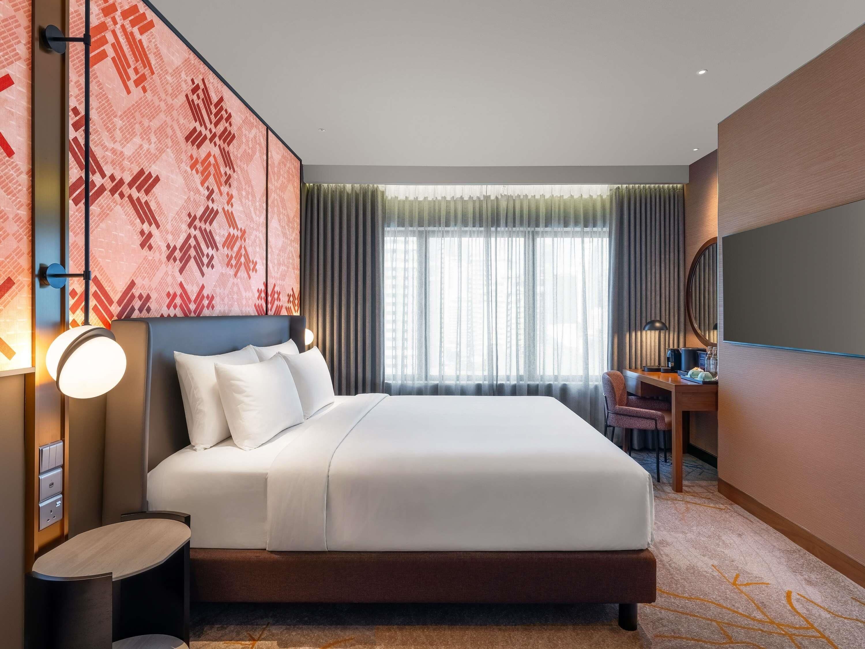 A modern hotel room with a large bed, a desk, and a view of the city through sheer curtains.