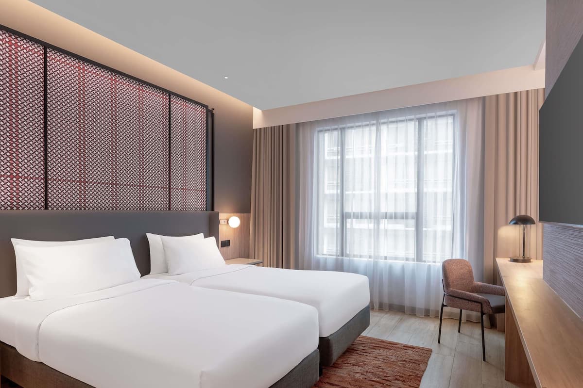 A hotel room with two beds, a desk, a chair, a large window with curtains, and a decorative wall panel.