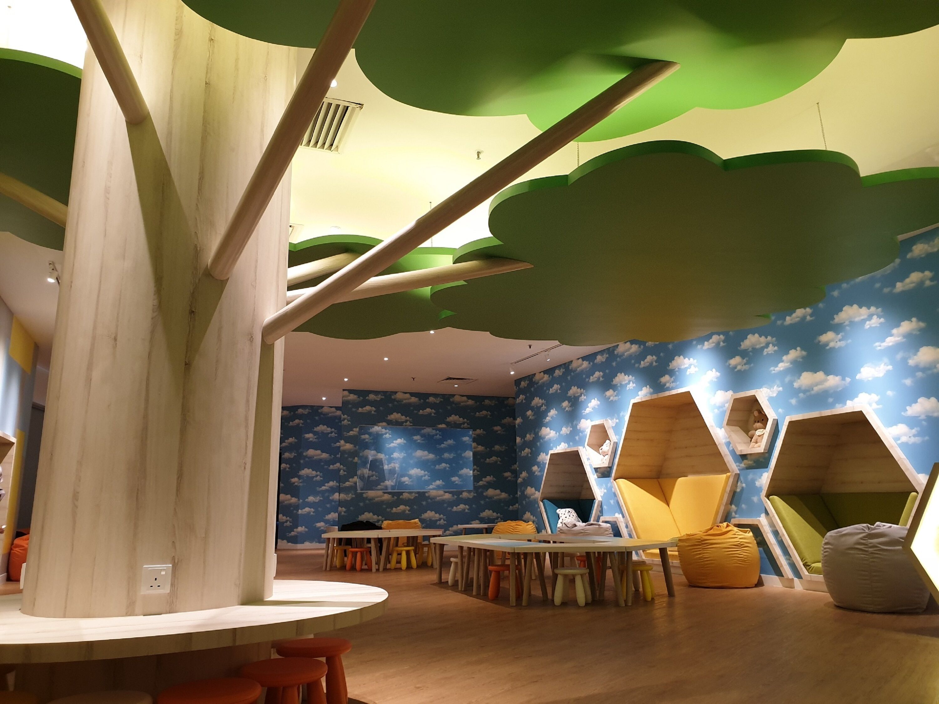 Zona infantil con una estructura de árbol, mesas, sillas y paredes con motivos de nubes.