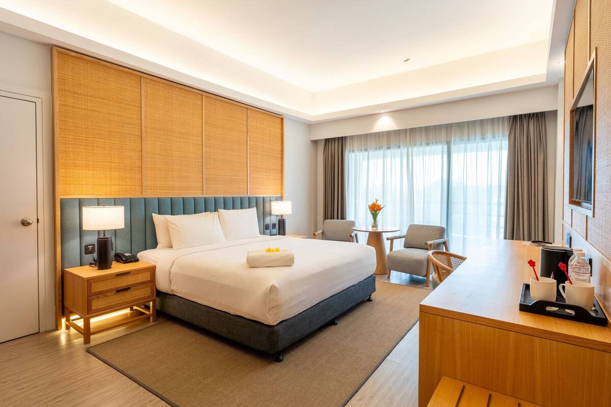 Ein Hotelzimmer mit einem Bett, Nachttischen, einem Schreibtisch, einem Sessel, einem kleinen Tisch mit einer Vase und einem großen Fenster mit Vorhängen.