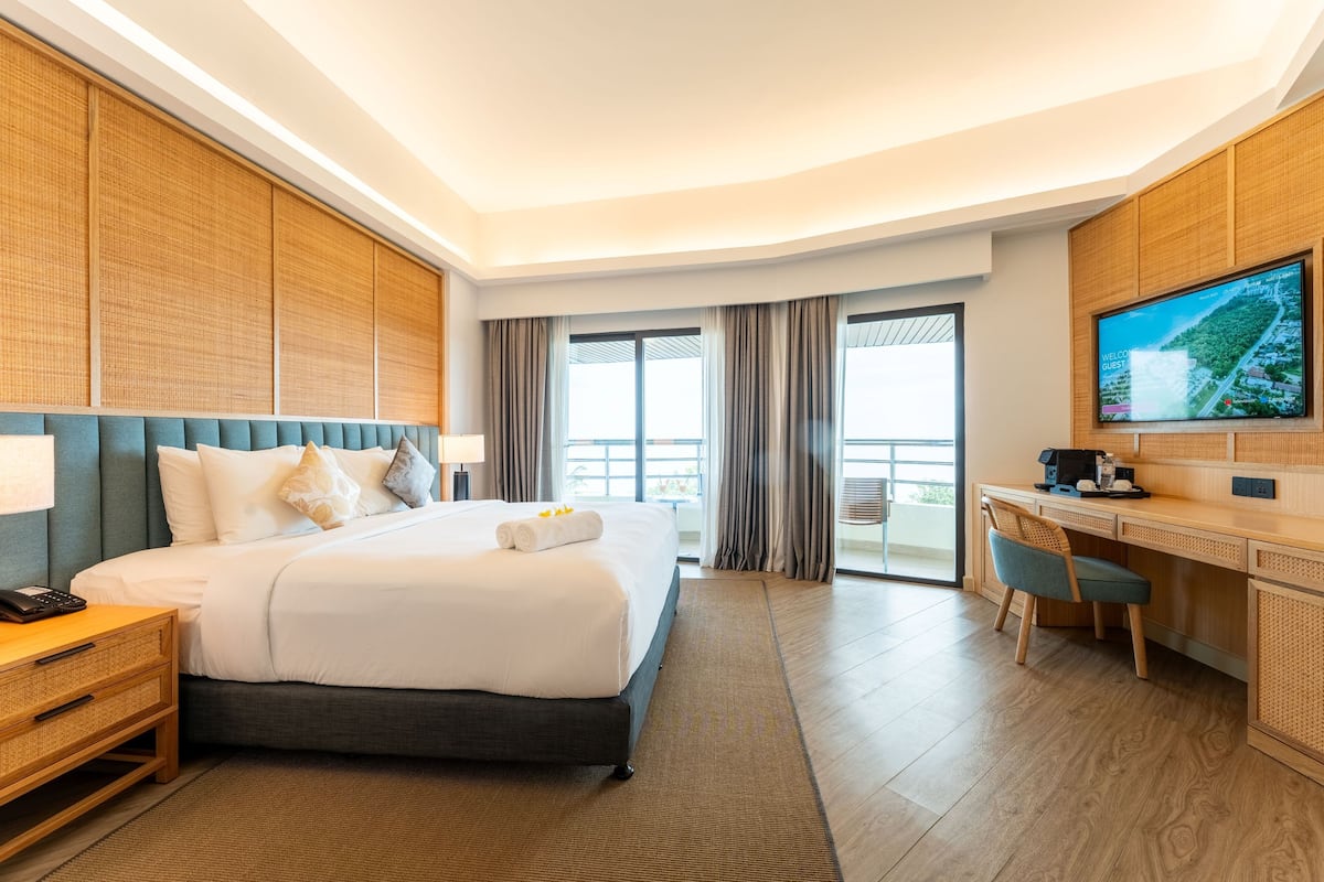 Ein Hotelzimmer mit einem großen Bett, einem Schreibtisch, einem Fernseher und einem Balkon mit Vorhängen.