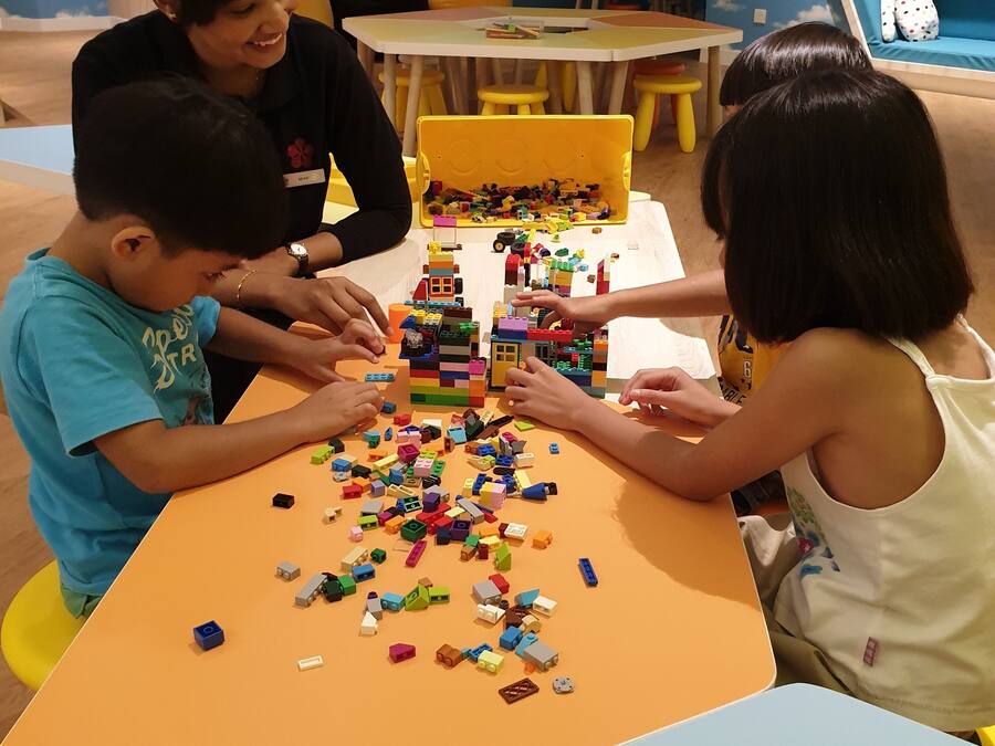 A group of people playing with building blocks at a table in a room with a blue wall and a cloud pattern.