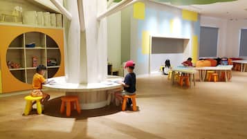 Un área de juegos infantiles con mesas redondas y taburetes naranjas, con un estante circular que exhibe juguetes y libros.