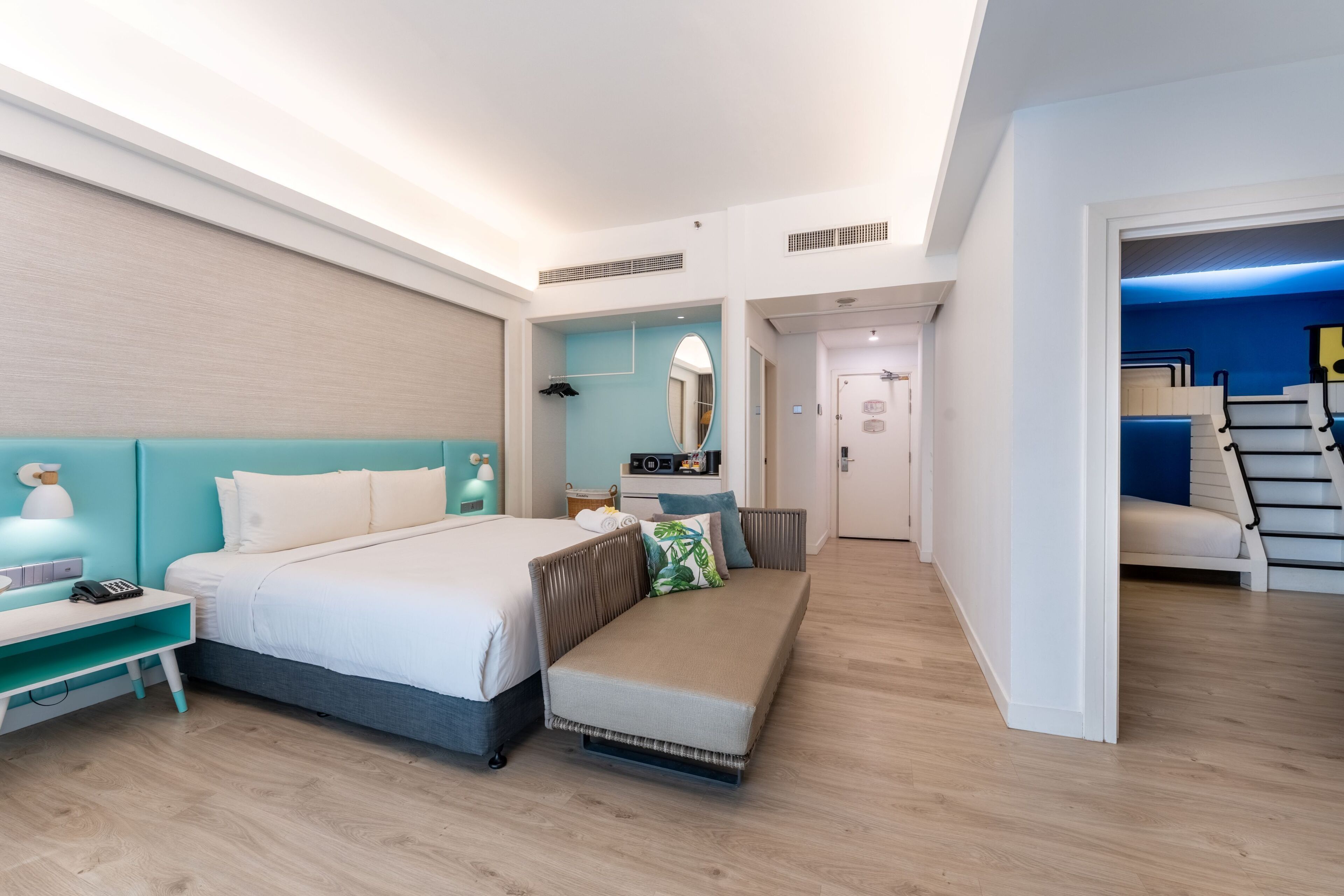 A modern hotel room with a bed, bedside table, and a bench. There is a mirror, a dresser, and a staircase leading to an upper level.