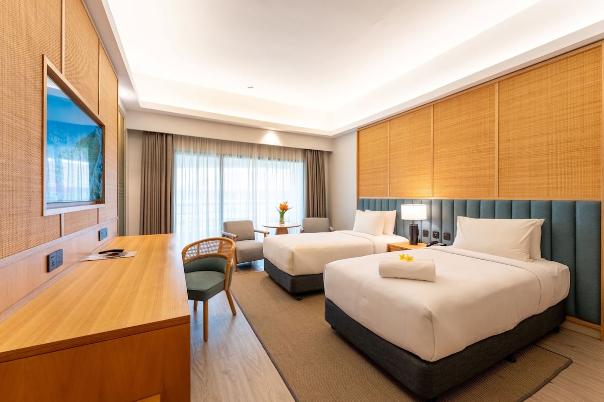 Ein Hotelzimmer mit zwei Betten, einem Schreibtisch, einem Stuhl, einem kleinen Tisch mit einer Vase und einem großen Fenster mit Vorhängen.