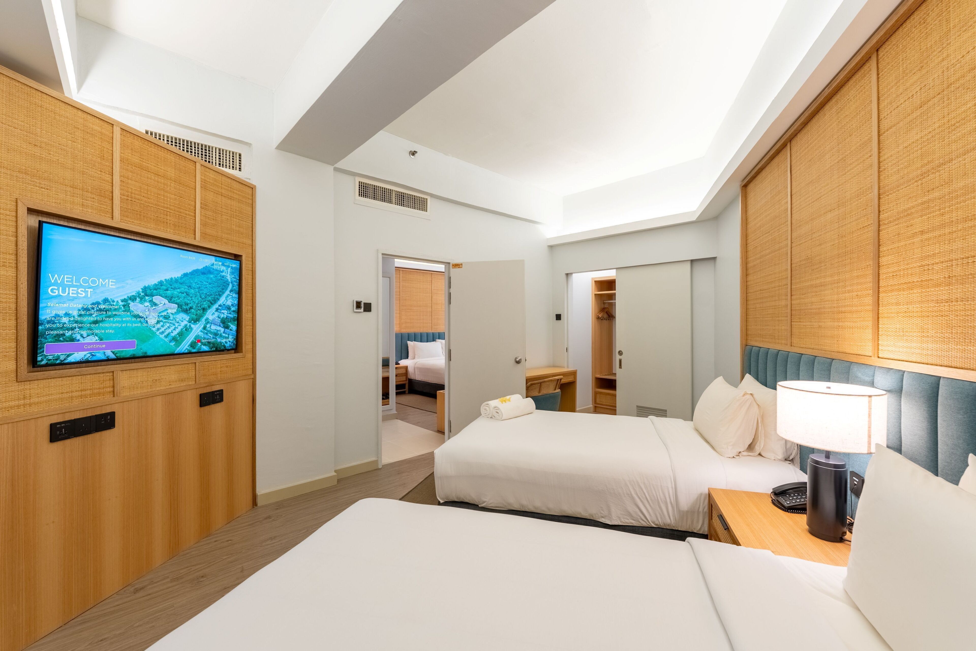 A hotel room with a television displaying a welcome message, two beds, a desk, and a lamp.
