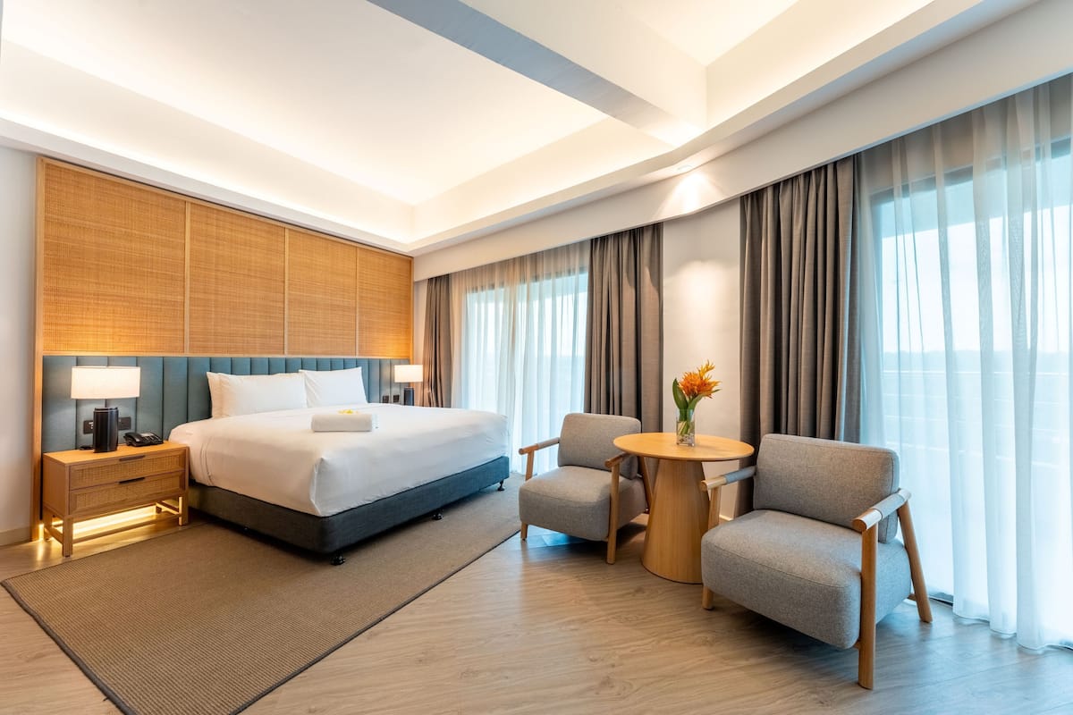 Ein modernes Hotelzimmer mit einem großen Bett, Nachttischen, einem kleinen runden Tisch mit Stühlen und Blick ins Freie durch große Fenster.