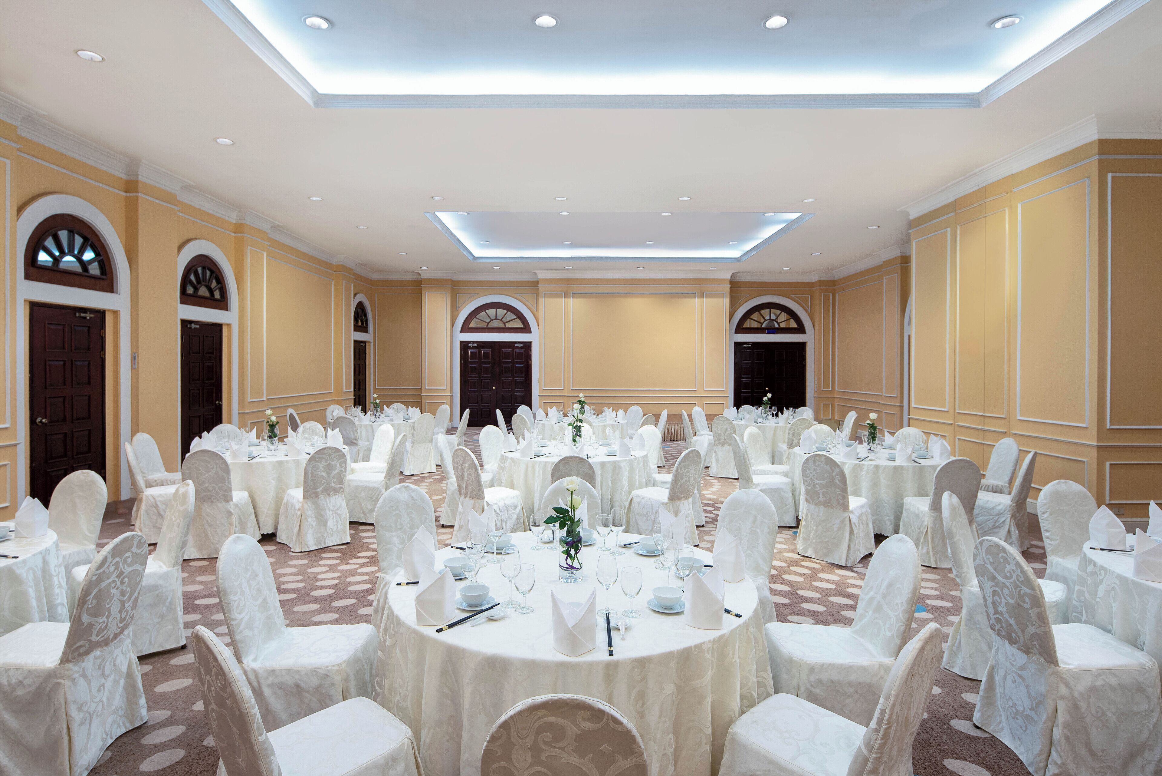 Een ruime balzaal met ronde tafels en witte stoelen, met een hoog plafond, inbouwverlichting en decoratieve lijsten.