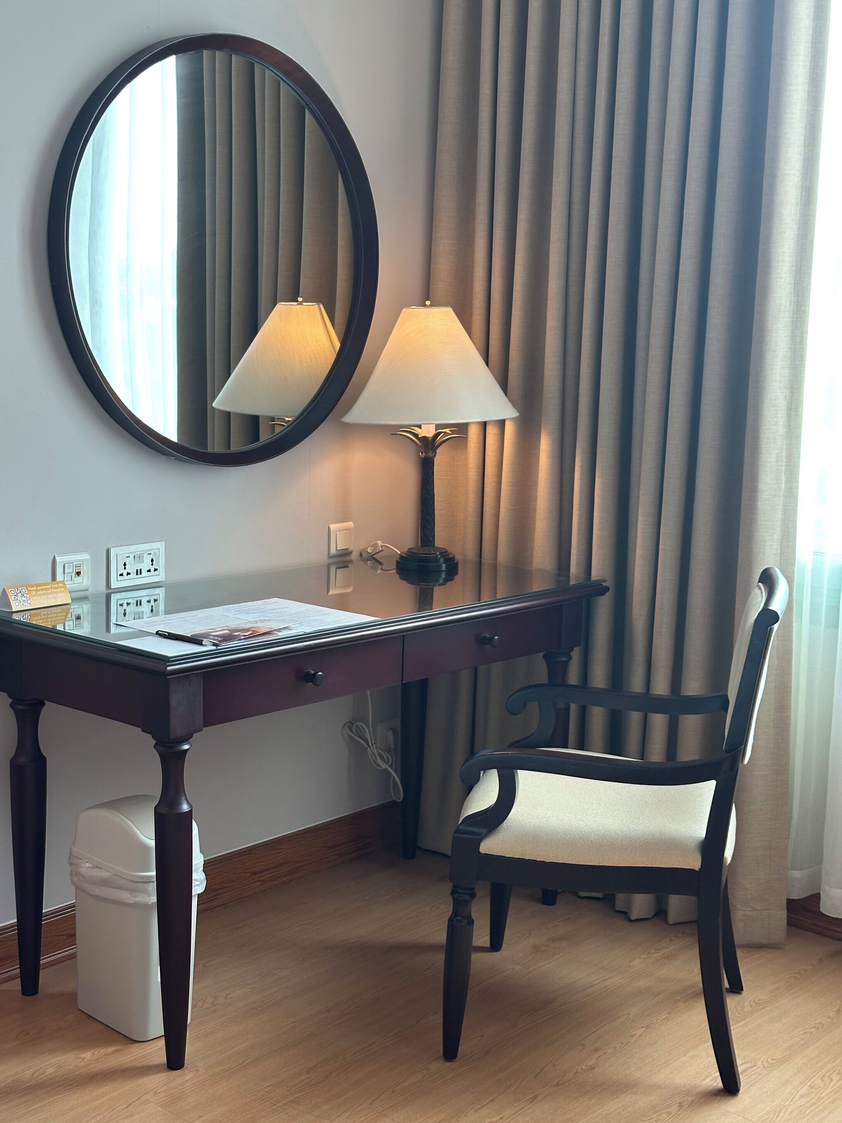 Habitación de hotel con un escritorio de madera, una silla, una lámpara, un espejo y una ventana con cortinas.