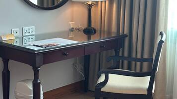 Habitación de hotel con un escritorio de madera, una silla, una lámpara, un espejo y una ventana con cortinas.