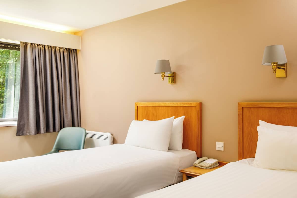 A hotel room with two beds, a chair, a bedside table with a phone, and a window with curtains.