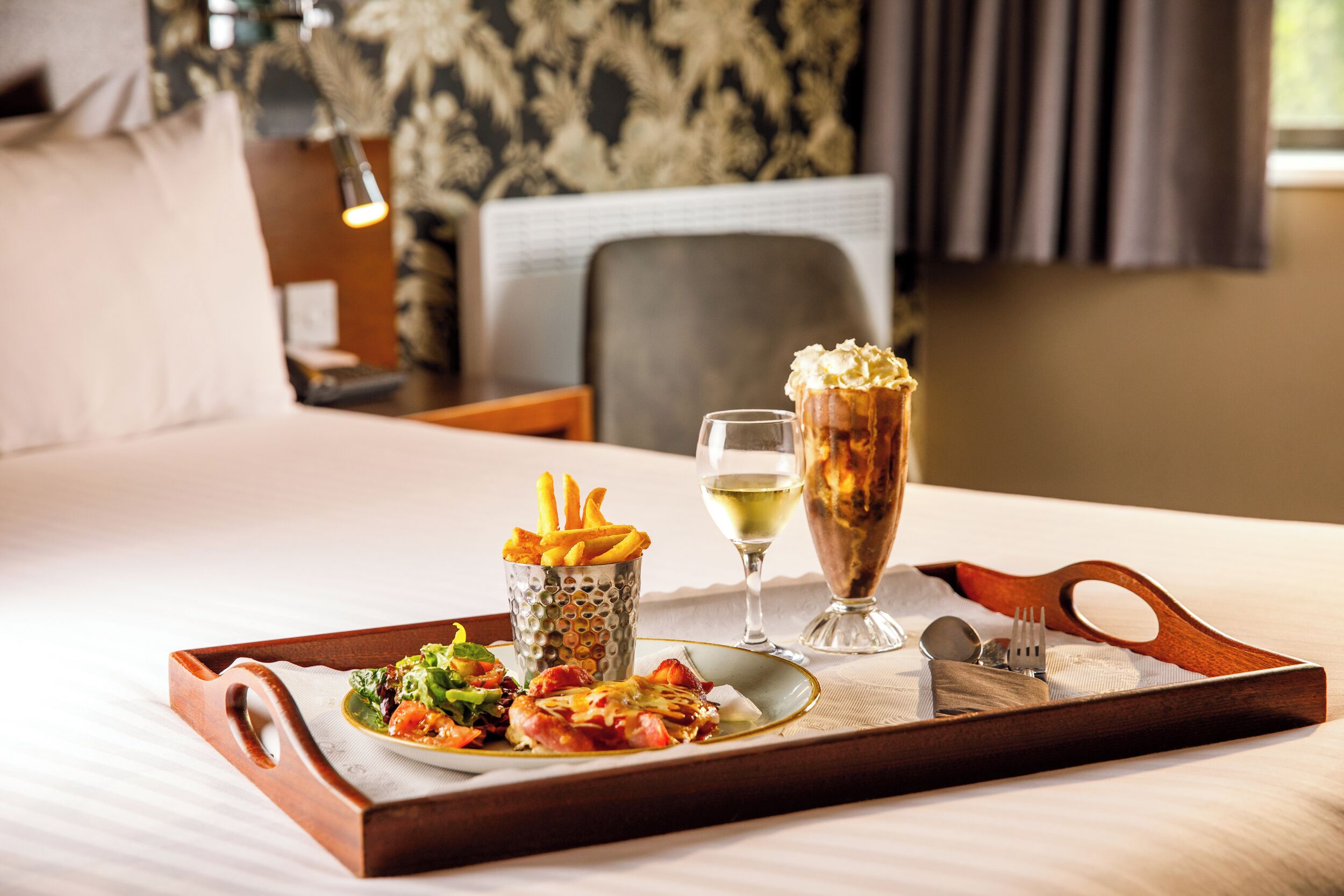 A tray with a plate of food, a glass of white wine, and a dessert on a bed in a hotel room.