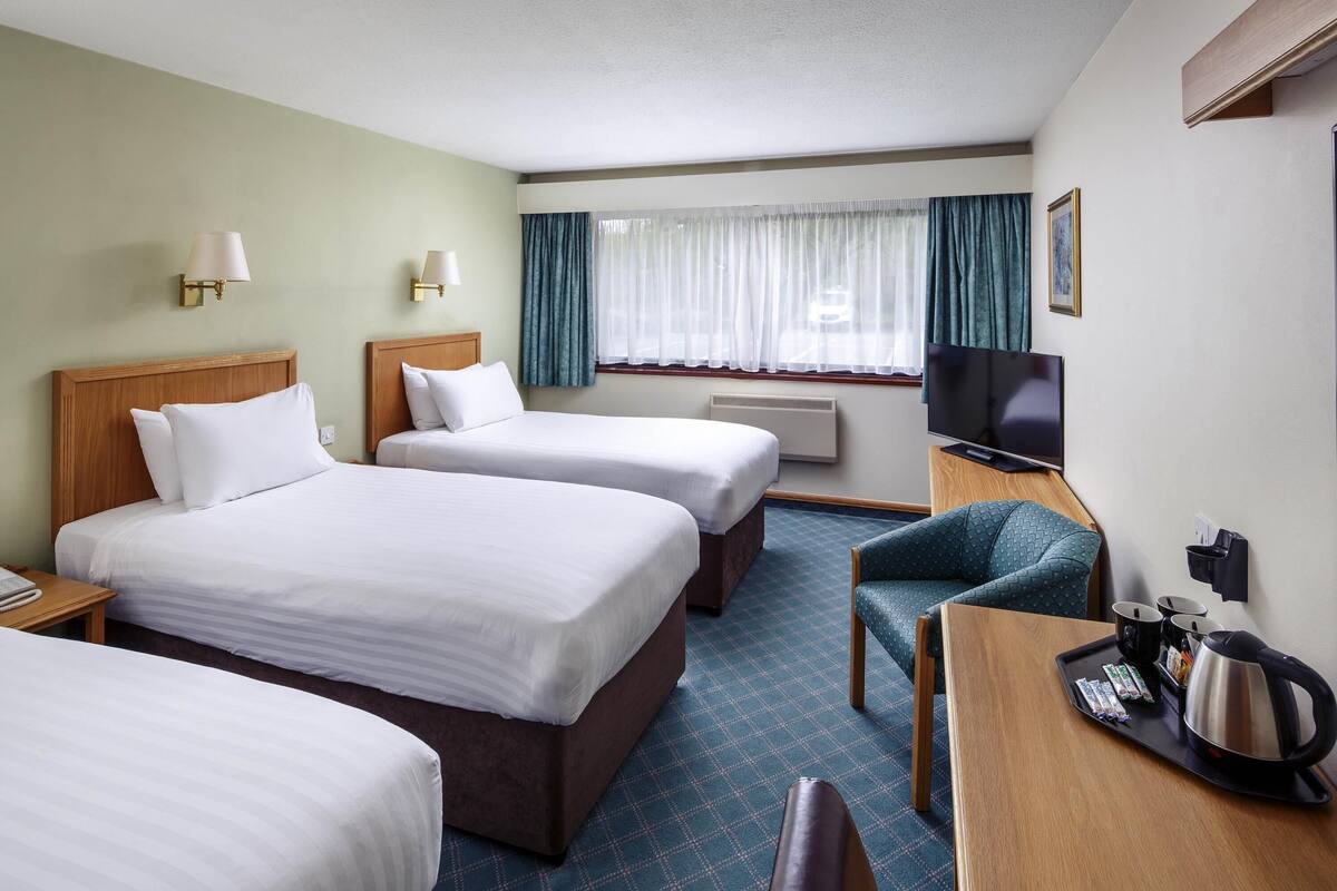 A hotel room with three beds, a desk with a kettle, a chair, a TV, and a window with curtains.