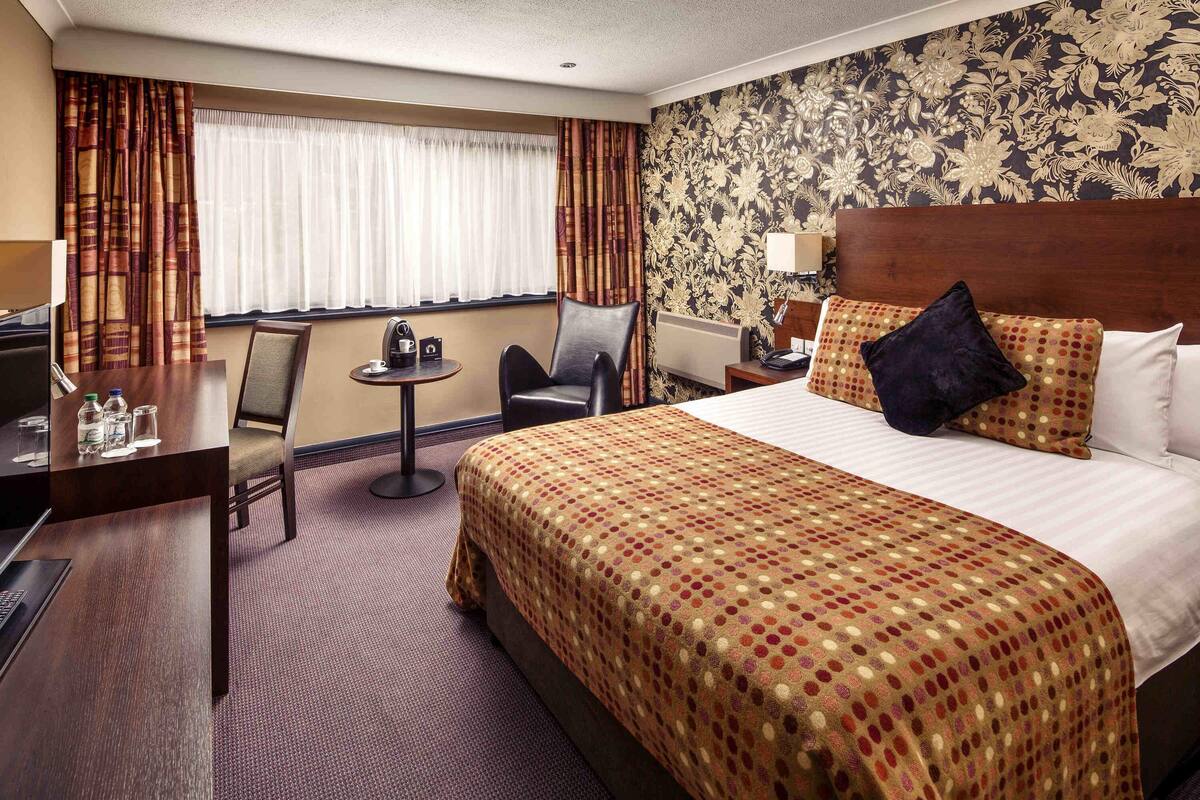 A hotel room with a large bed, a desk, a chair, a small table with a lamp, and a window with curtains.