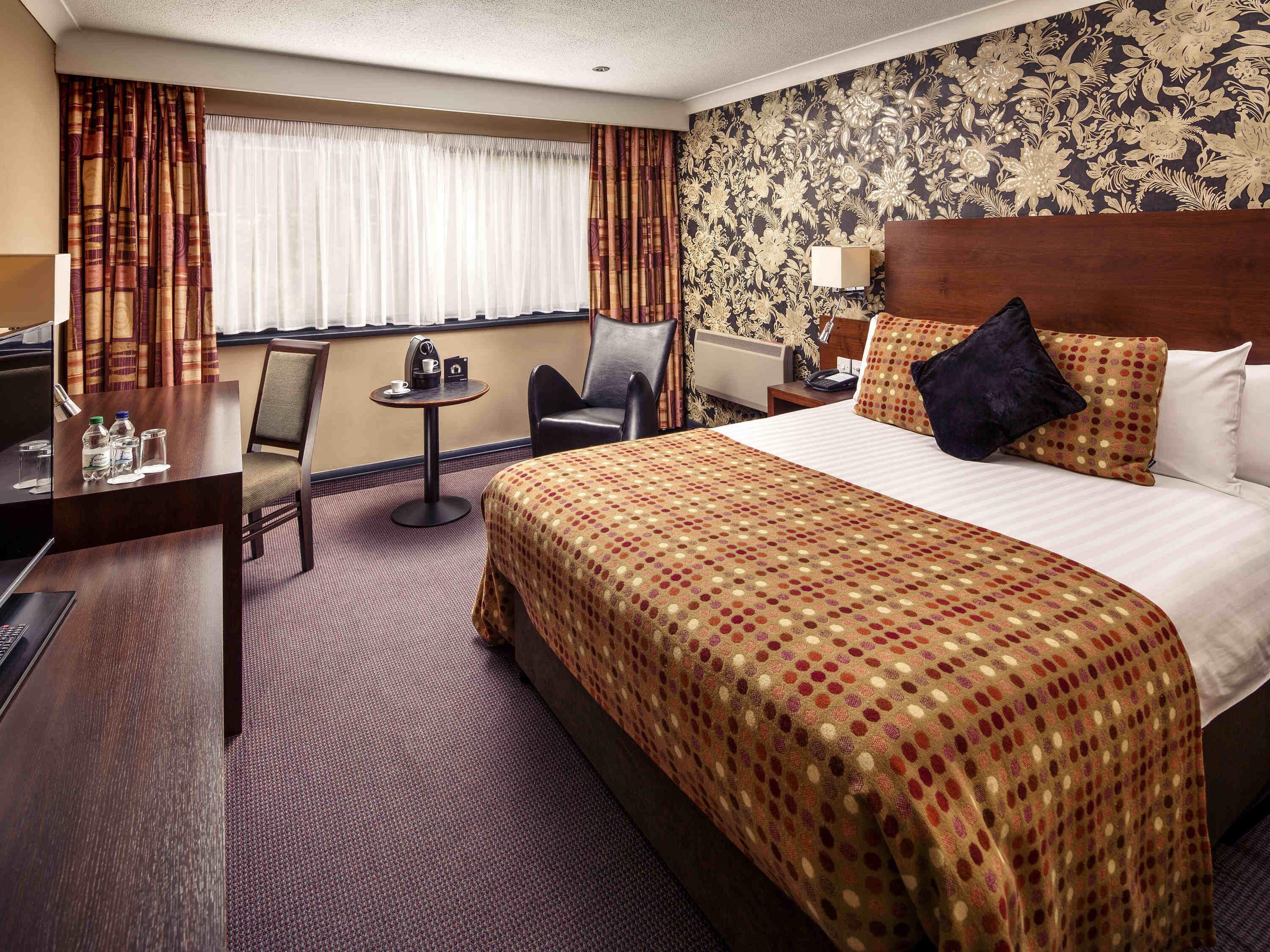 A hotel room with a large bed, a desk, a chair, a small table with a lamp, and a window with curtains.