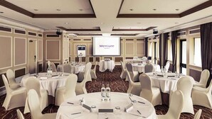 Un salón de conferencias con mesas redondas, sillas blancas y una pantalla de proyección en un hotel.
