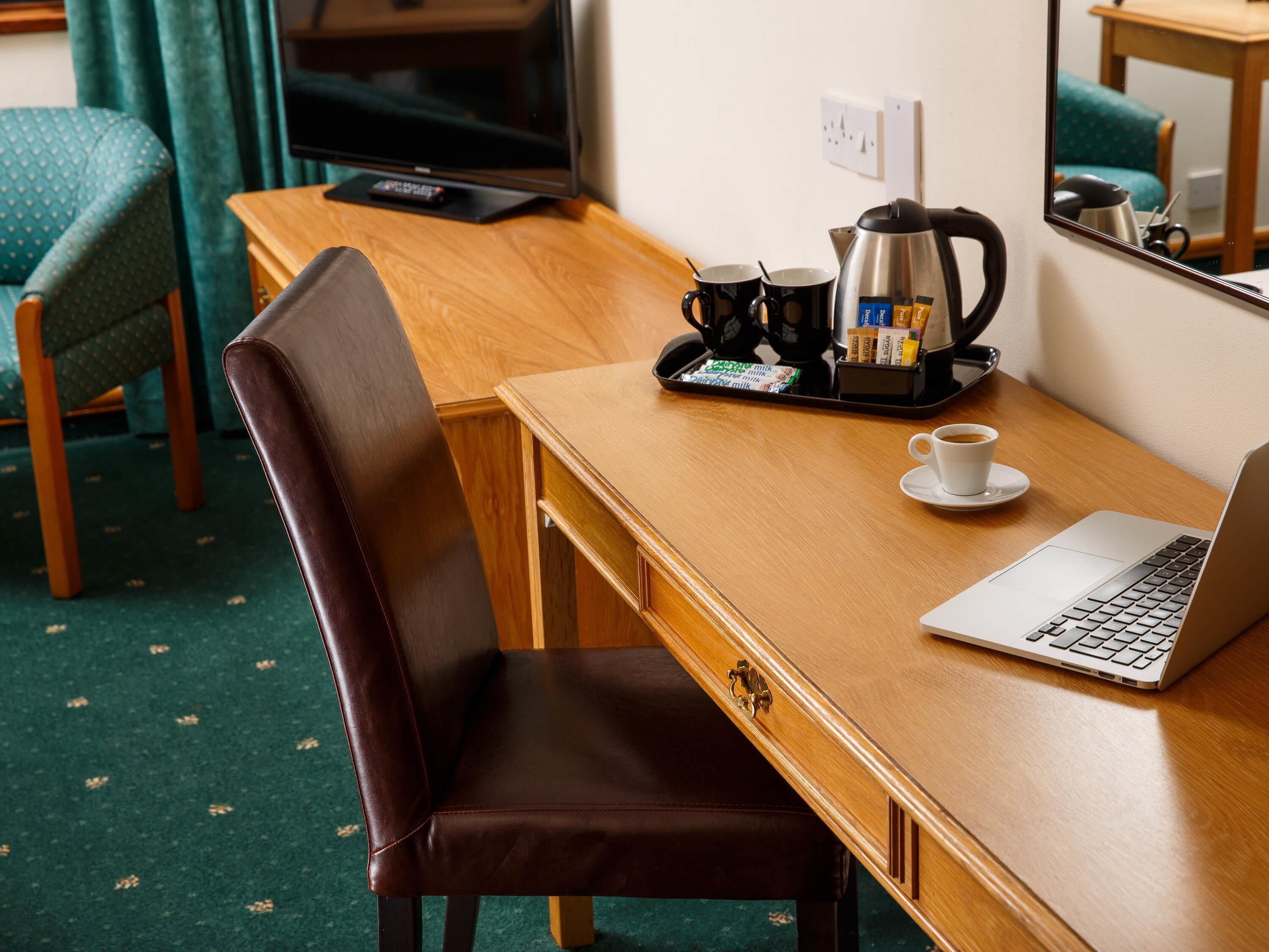 A hotel room with a wooden desk, a laptop, a coffee maker, and a tray with coffee cups and milk packets.