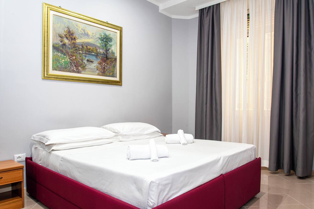 A neatly made bed with white linens and red headboard, a painting on the wall, and a window with grey curtains.