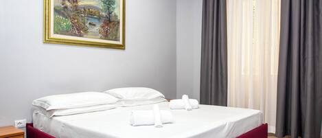 A neatly made bed with white linens and red headboard, a painting on the wall, and a window with grey curtains.