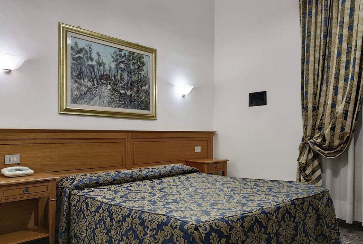 A hotel room with a wooden headboard, a bed with a patterned bedspread, a bedside table with a telephone, and a wall-mounted painting.