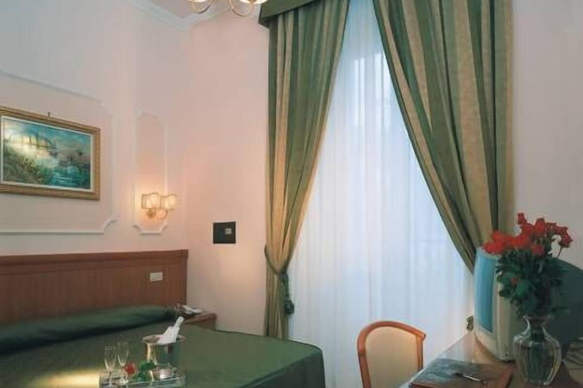 A hotel room with a bed, a desk with a vase of flowers, a chair, a painting on the wall, and a window with curtains.