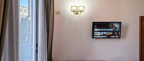 Una habitación de hotel con un escritorio, un televisor en la pared mostrando la interfaz de un servicio de streaming, y una ventana con cortinas.