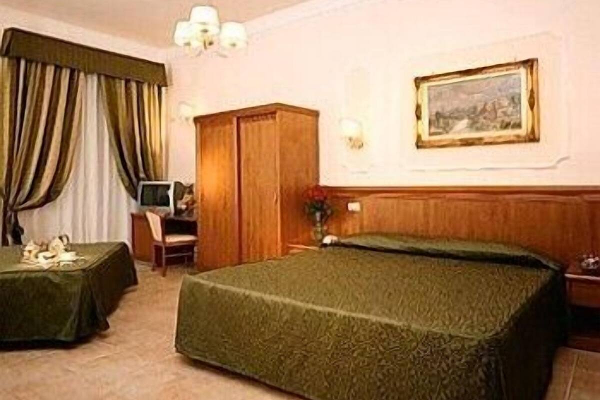 A hotel room with a bed, a desk with a chair, a wardrobe, and a painting on the wall.